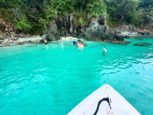Guests wading and snorkeling in a turquoise cove below rocky cliffs, seen from a boat bow, St John USVI